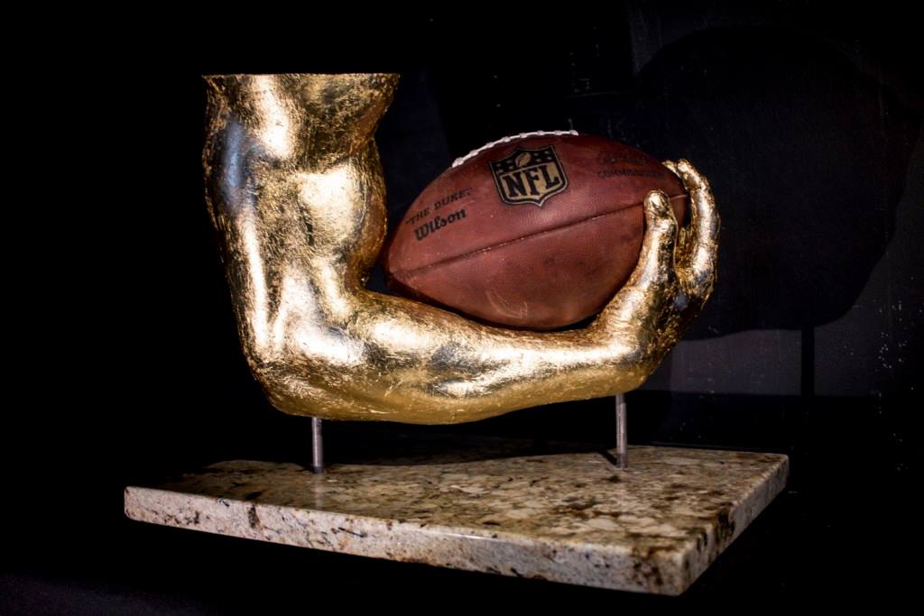 A custom-casted gold arm and hand sculpture holding an official NFL football, mounted on a polished stone base. The intricate detailing captures every curve and muscle, preserving the essence of the athlete’s grip.